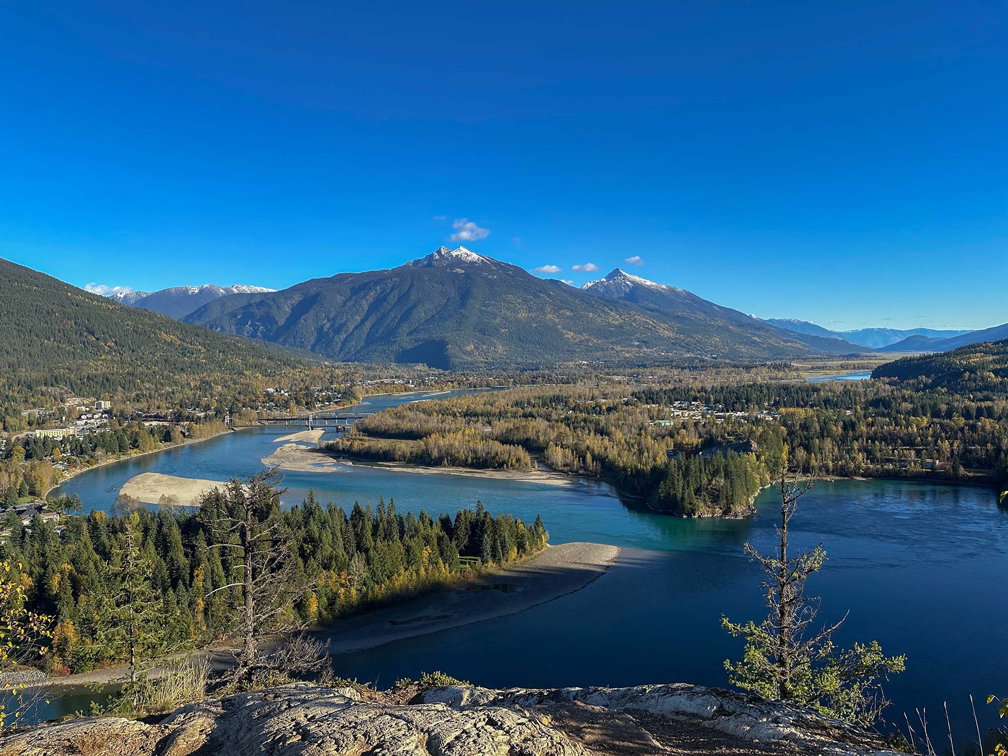 Panoramic view of a winding blue river, forested valley, bridge, town, and snow-capped mountains.