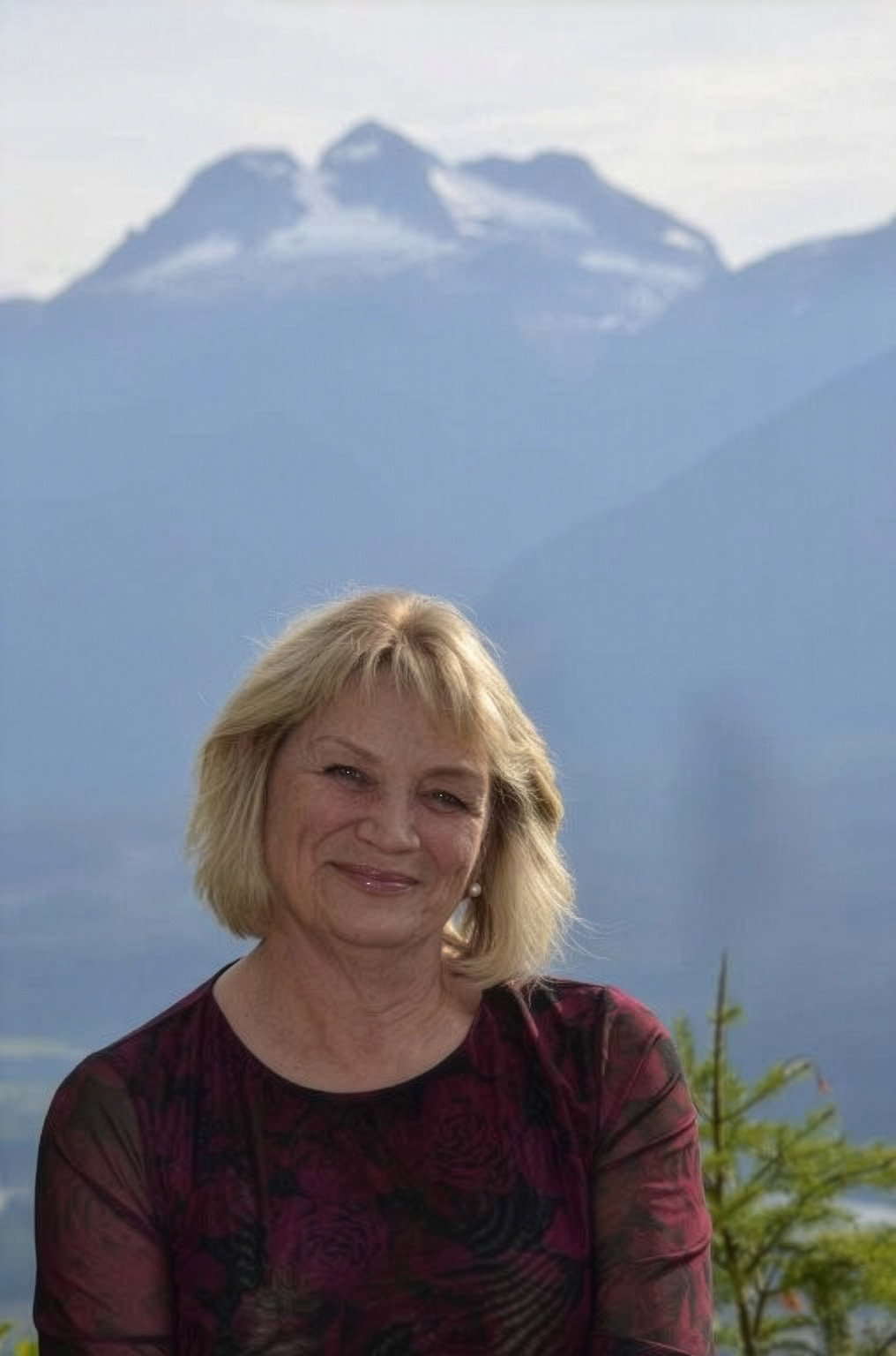 Smiling blonde woman in a burgundy patterned top poses before hazy blue snow-capped mountains and greenery.