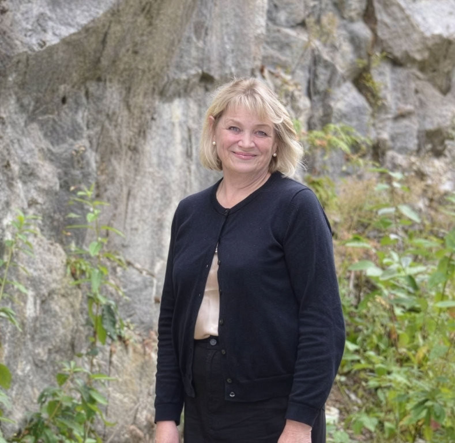 Smiling blonde woman in a dark cardigan standing before a gray rock wall with green plants.
